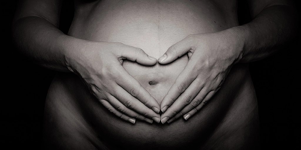 Black-and-white maternity detail, hands cradling the pregnancy bump in a heart shape, Barcelona.